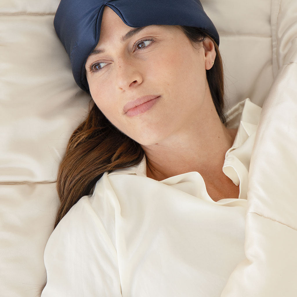 A woman wearing a dark blue sleep mask pushed up on her forehead lies in bed, covered with a cream-colored blanket, and gazes thoughtfully to the side. She wears a white shirt and has long brown hair.