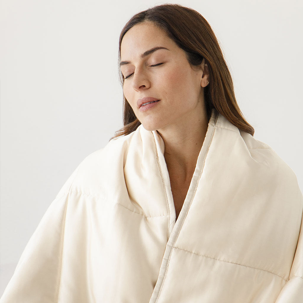 A woman with long brown hair, eyes closed, is wrapped in the drowsysleepco Dusty Gold Silk Weighted Blanket, looking calm and relaxed against a plain light background.