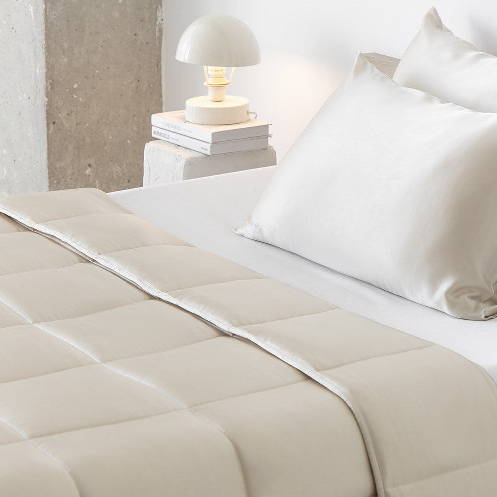 A neatly made bed features light cream bedding with a quilted comforter and the drowsysleepco Dusty Gold Silk Weighted Blanket. A small lamp on stacked books rests on a bedside table by a concrete wall, creating a bright, minimal look.
