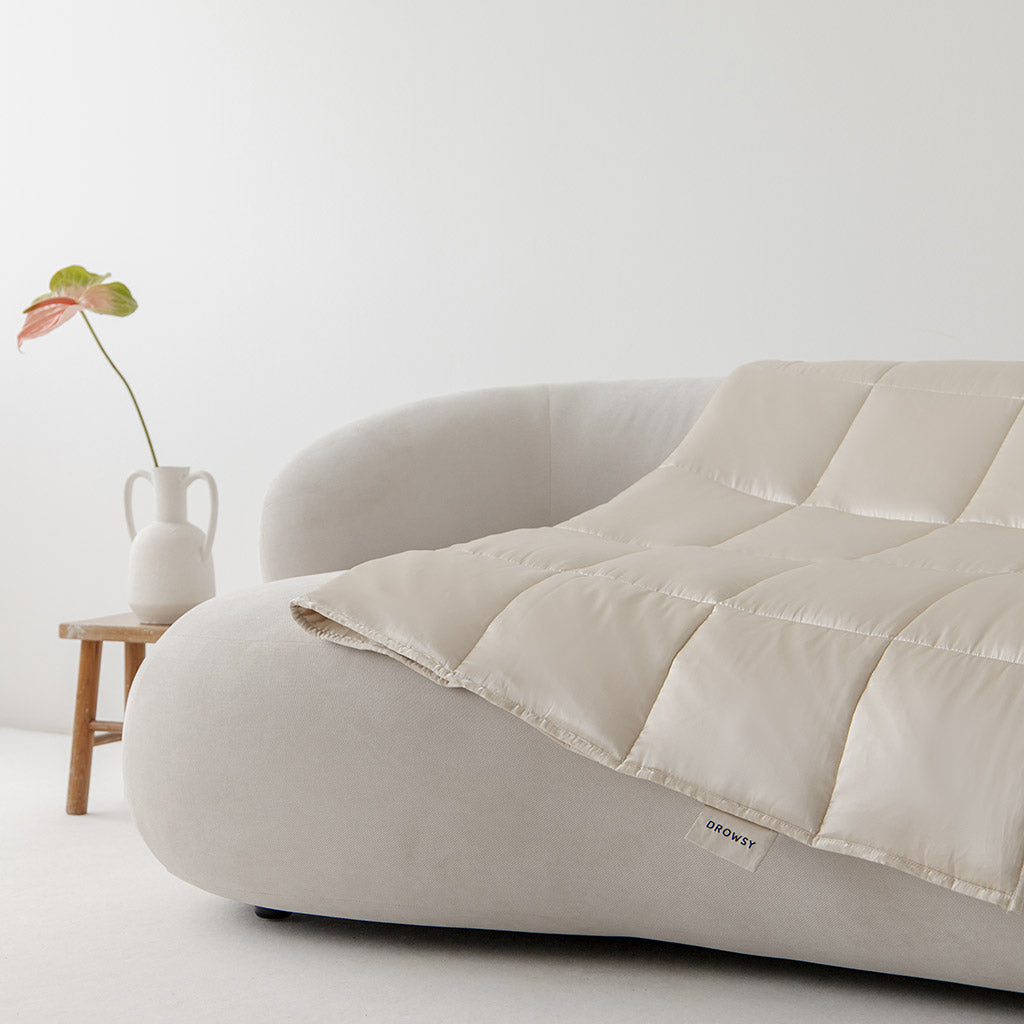 The drowsysleepco Dusty Gold Silk Weighted Blanket is draped over a modern curved white sofa, with a small wooden table in the background holding a white vase and a single green-pink leaf, creating a minimal, bright aesthetic.