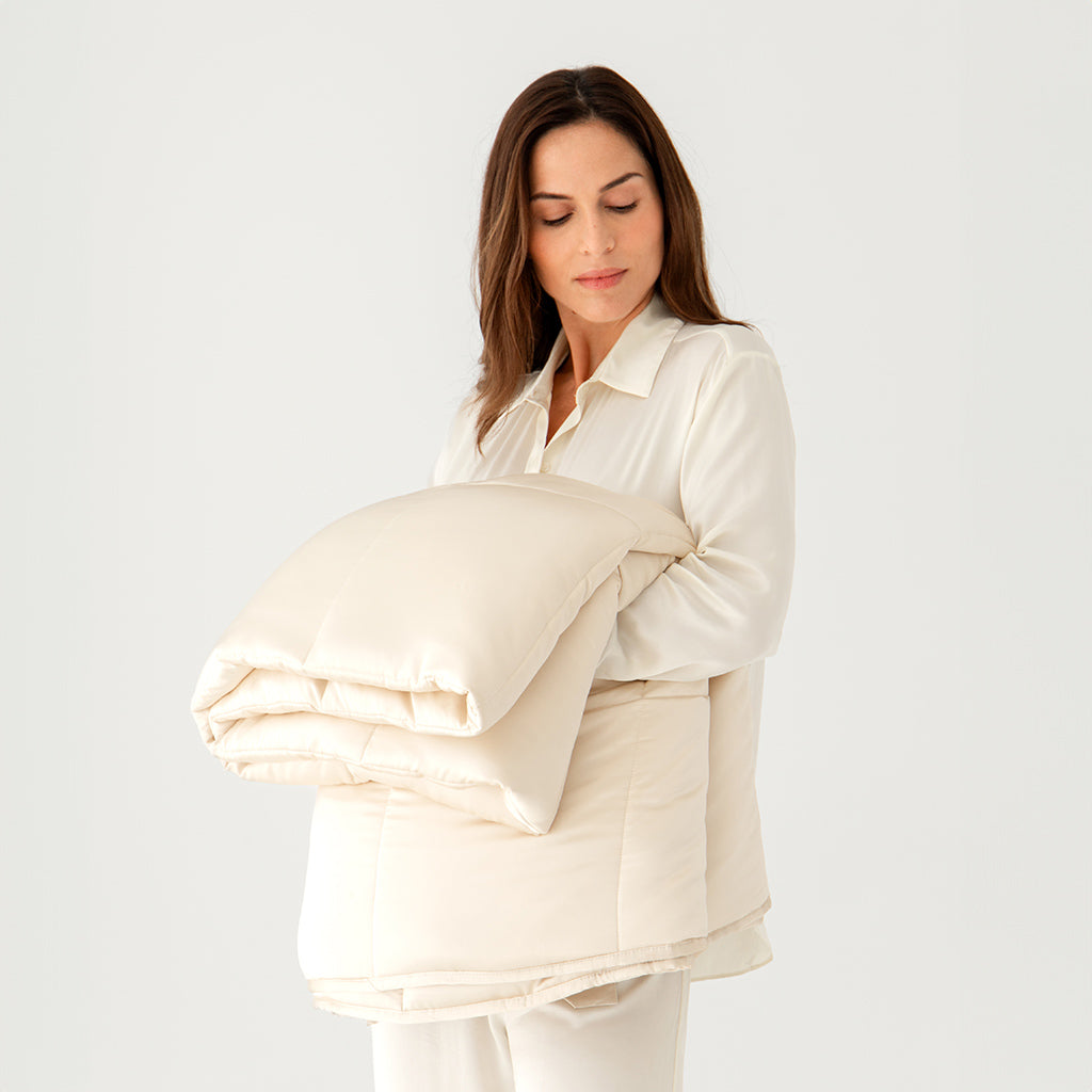 A woman in cream-colored pajamas holds a folded, light beige comforter against her chest, standing against a plain white background.