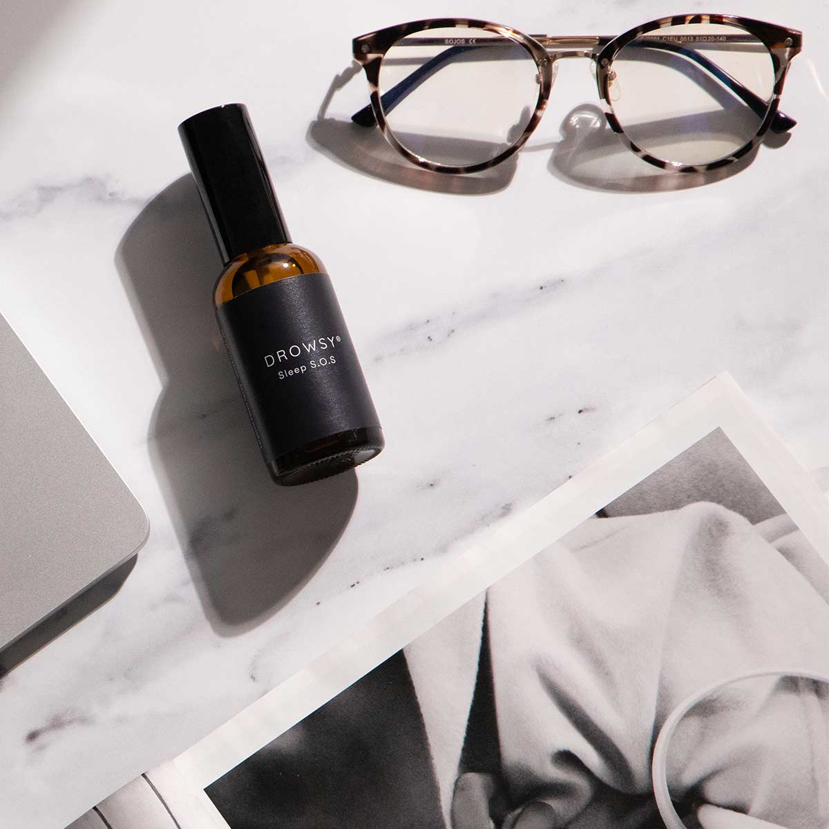A small brown bottle labeled Drowsy Sleep SOS sits on a marble surface next to tortoiseshell eyeglasses, a corner of a laptop, and a black-and-white magazine page.