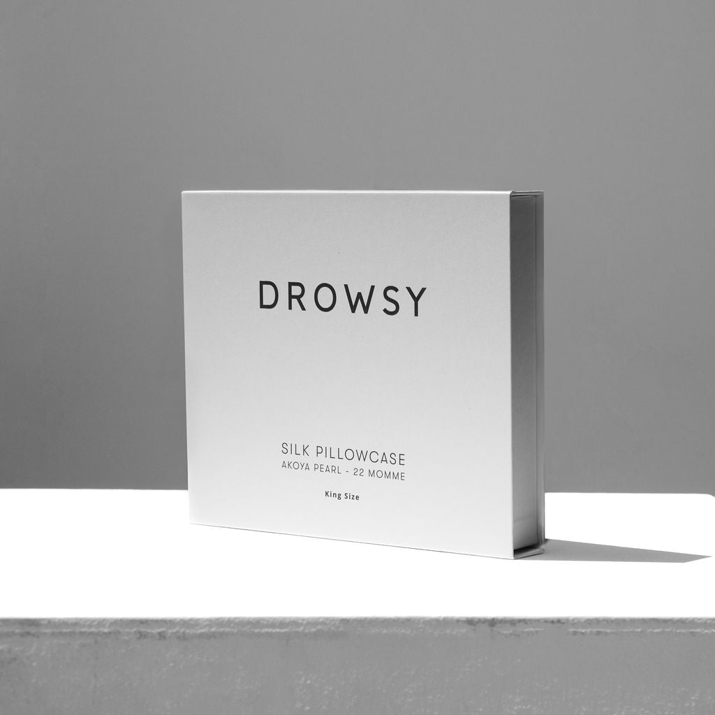 A minimalist photo of a white box labeled drowsysleepco containing a Rajah Cushion Cover. The box features simple black text and is displayed on a plain white surface with a gray background.