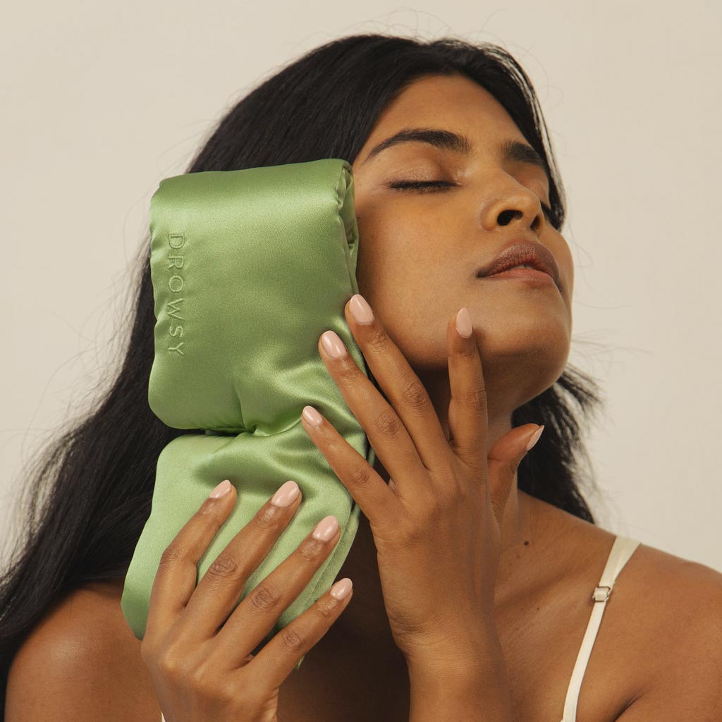 A woman with long dark hair, eyes closed in relaxation, holds the Pistachio Green sleep mask by drowsysleepco to her face. She wears a cream top and stands against a neutral background, embodying the comfort of beauty sleep.
