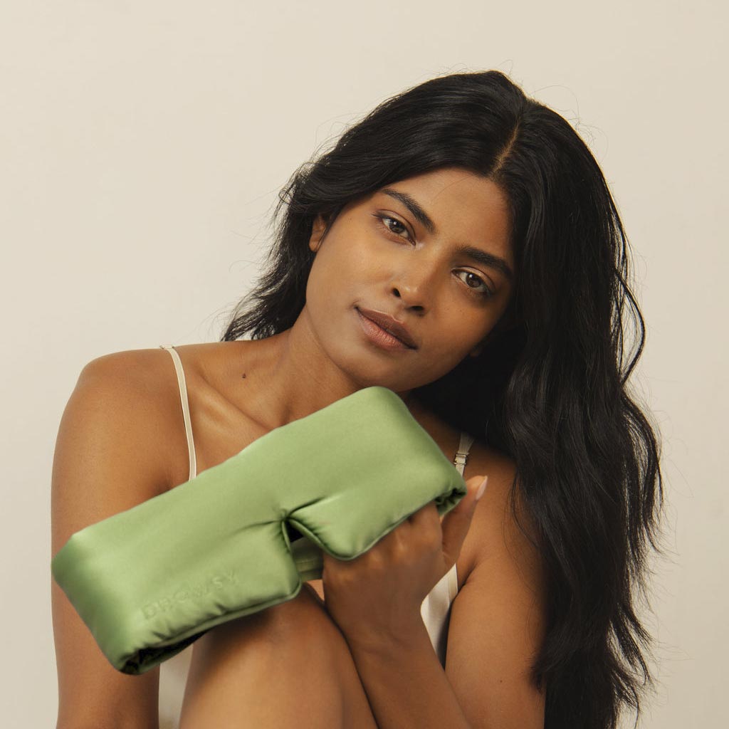 A woman with long dark hair in a white tank top holds the drowsysleepco Pistachio Green silk sleep mask to her chest, looking at the camera against a plain light background.