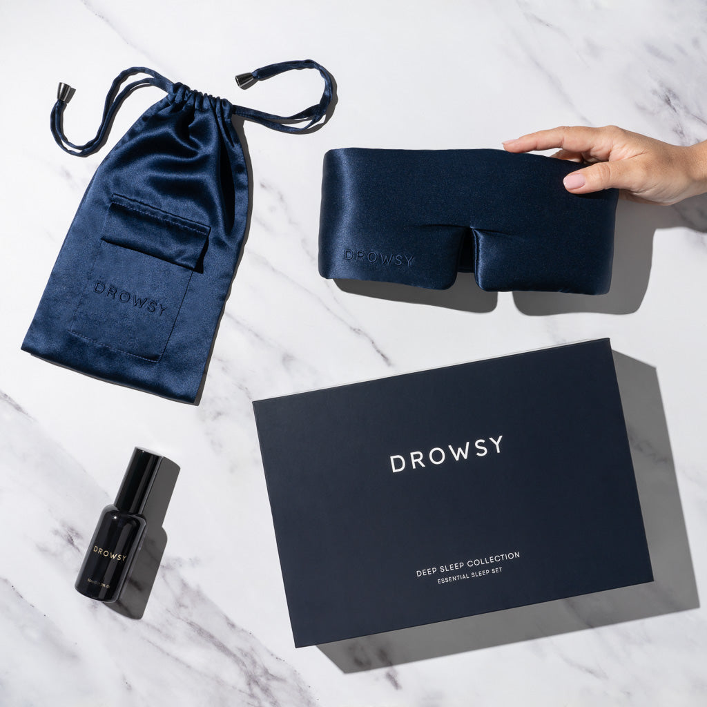 A hand holds the Midnight Blue Deep Sleep Collection by drowsysleepco: a navy silk sleep mask, matching pouch, and deep sleep pillow spray, all displayed with a dark blue box on white marble.
