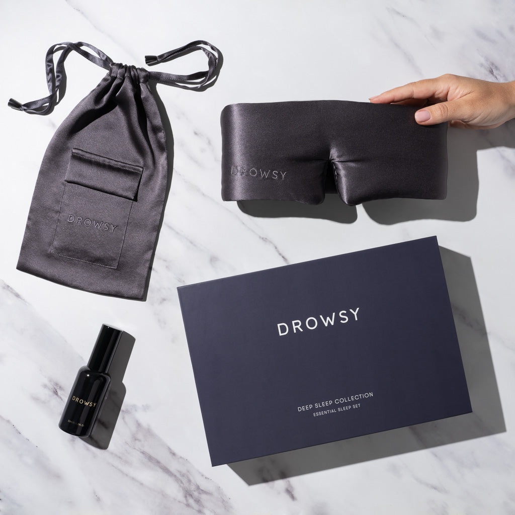 A hand holds a luxury silk sleep mask on a marble surface with sleep essentials: a black drawstring pouch, deep sleep pillow spray, and the drowsysleepco Moonlight Shadow Deep Sleep Collection dark blue box.