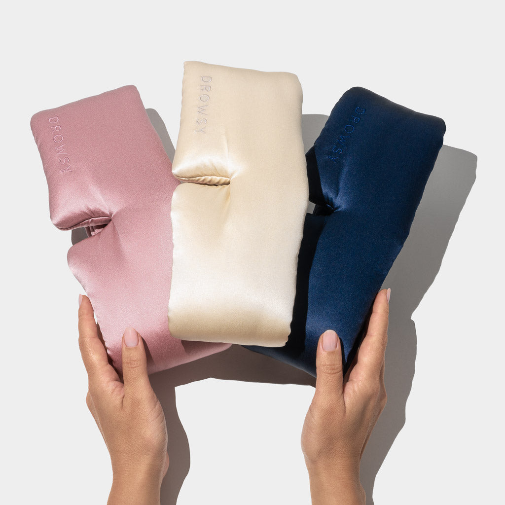 Two hands hold drowsysleepco’s Besties Pack: three luxury mulberry silk sleep masks in pink, cream, and navy blue, side by side on a white background. Each padded blackout eye mask offers ultimate comfort.
