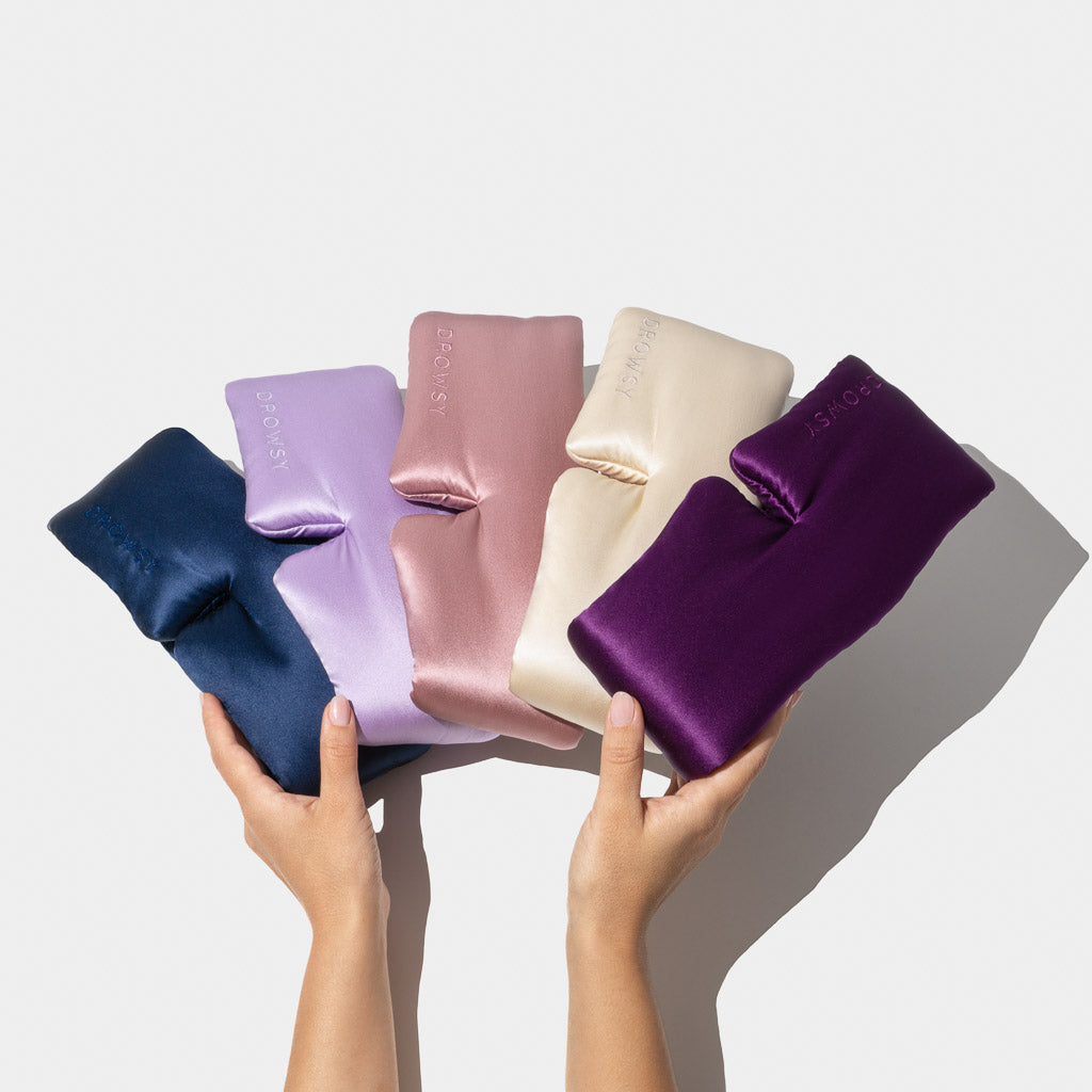Two hands hold the drowsysleepco Family Pack: five luxury silk masks in navy blue, lavender, pink, champagne, and purple, fanned out against a plain background—perfect for stylish beauty sleep.