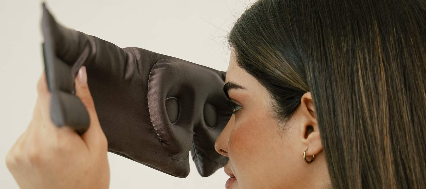 A woman with straight brown hair and gold hoop earrings holds a large, padded black eye mask up to her face, preparing to wear it. The background is neutral and out of focus.
