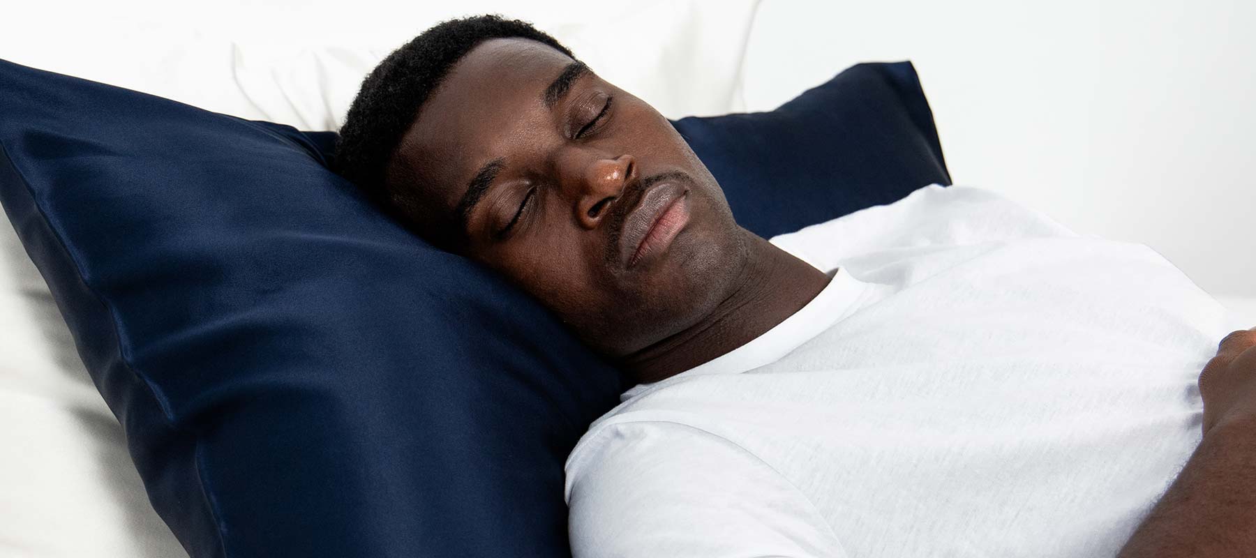 A man in a white shirt sleeps peacefully on a drowsysleepco Midnight ritual bundle pillowcase in Midnight Blue, surrounded by all-white bedding for a calm atmosphere.