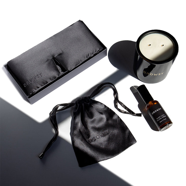 A black silk eye mask, black drawstring pouch, small brown bottle, and black candle with two wicks are arranged neatly on a white surface in bright light. The items are branded Drowsy.