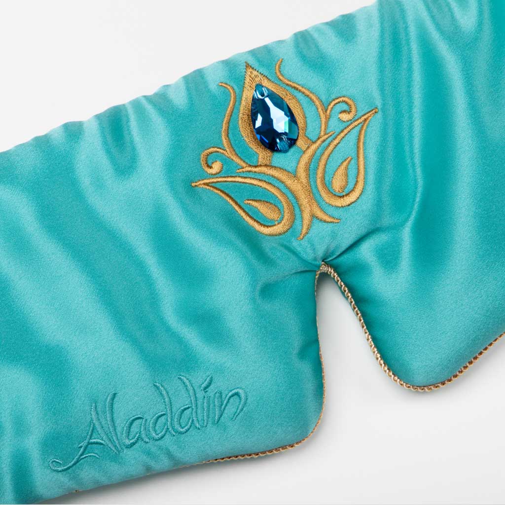 The Jasmine Jewel Sleep Mask by drowsysleepco features turquoise mulberry silk, gold embroidery, a large blue gem, and “Aladdin” in cursive on the left side for a shiny, luxurious blackout effect.