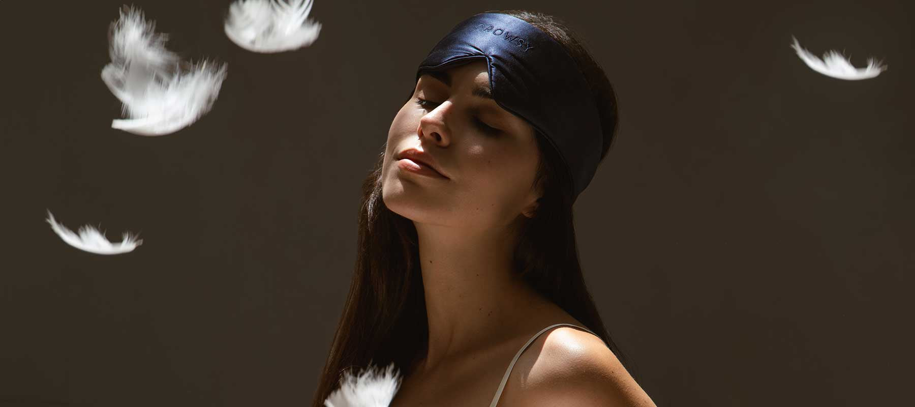 A woman wearing a dark sleep mask tilts her head back with a peaceful expression as white feathers float around her against a dark background.