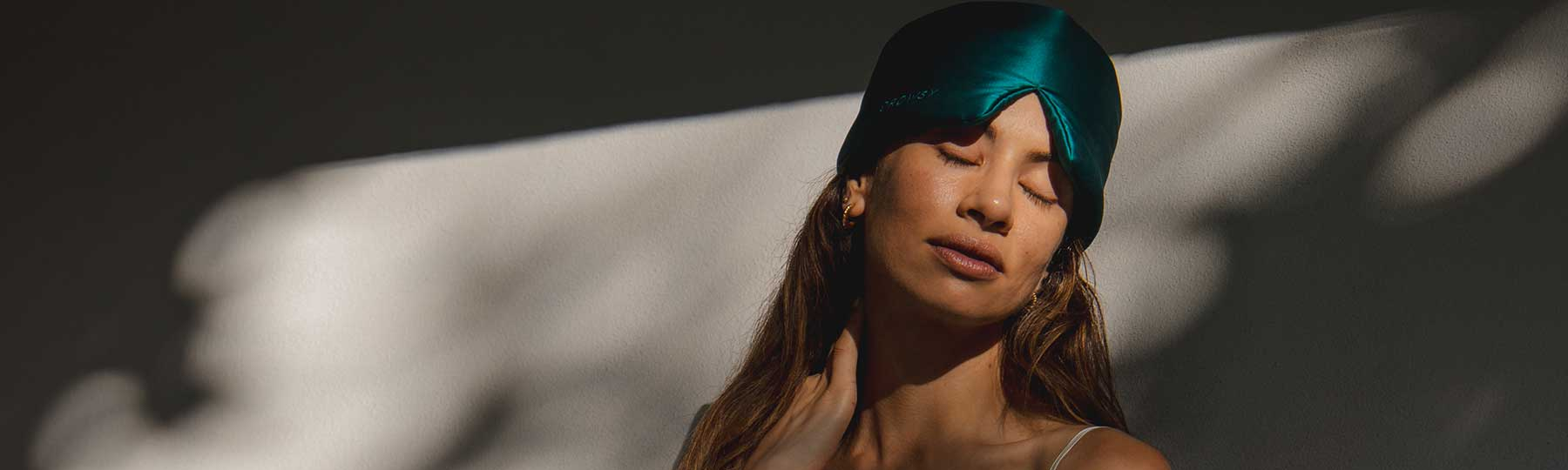 A woman with long hair, wearing a dark green satin sleep cap and a white strap top, stands with her eyes closed, gently touching her neck, bathed in soft sunlight with shadows on the wall behind her.