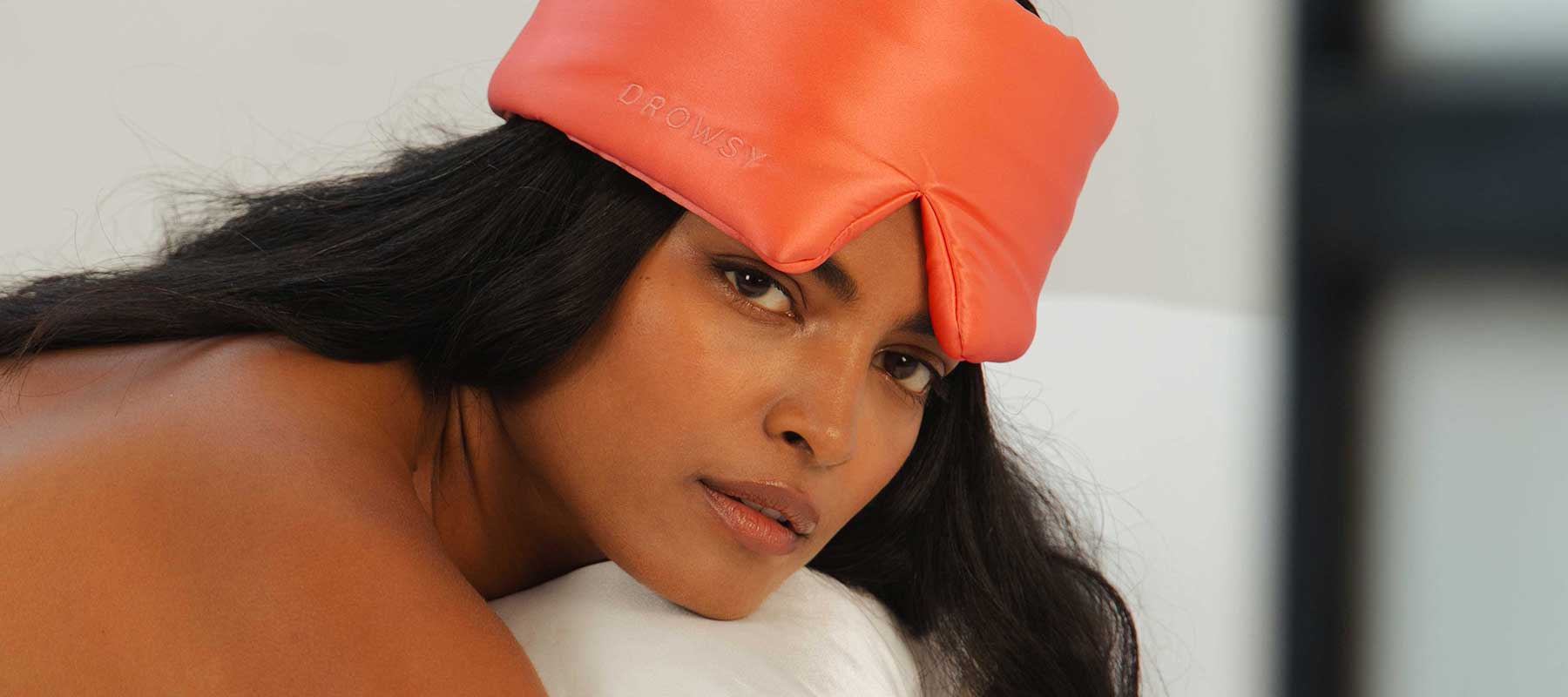 A woman with long dark hair lies on her side, wearing a coral-colored satin sleep mask on her forehead. She gazes softly at the camera against a neutral, softly-lit background.