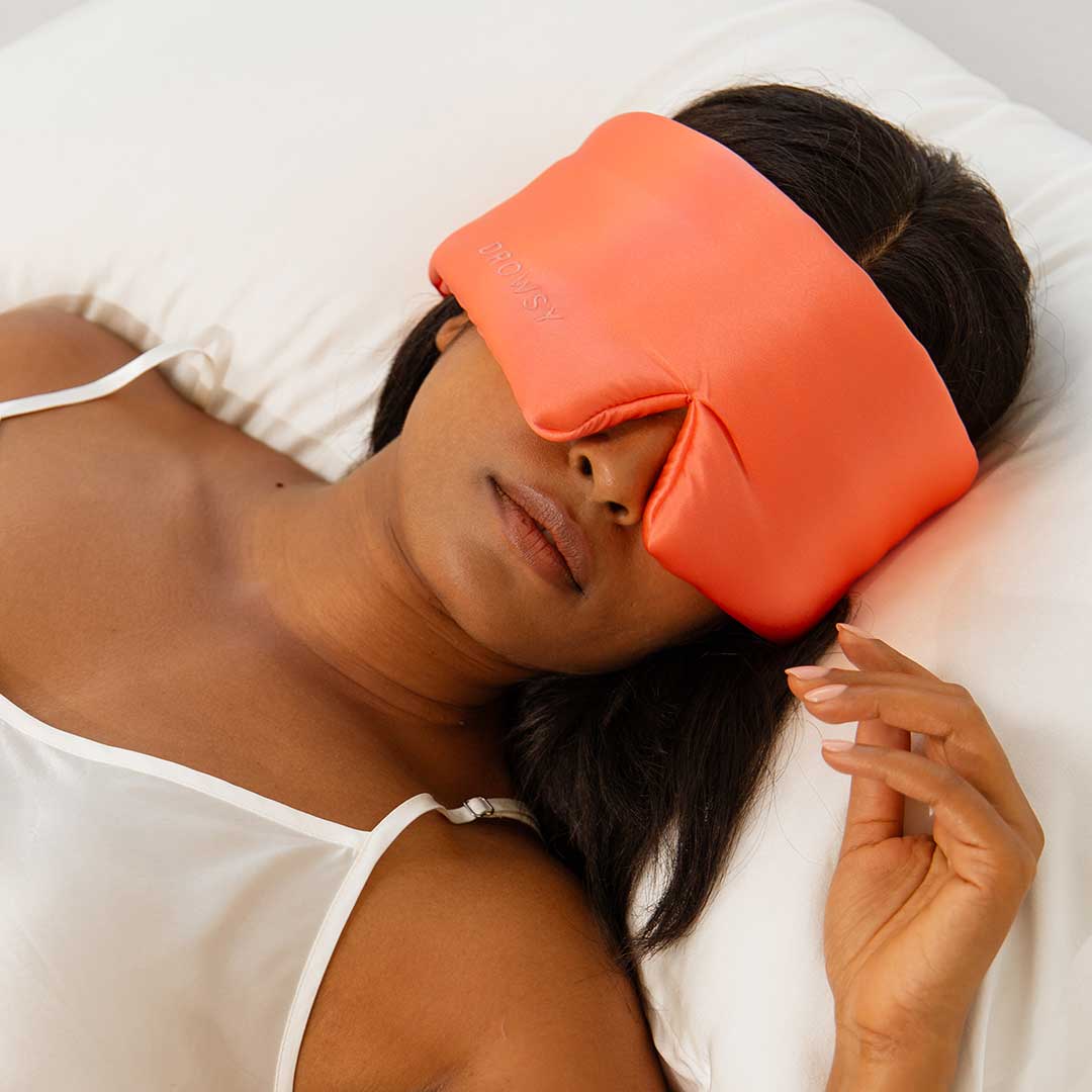 A woman rests peacefully on a white pillow, wearing a coral silk Sleep Mask by drowsysleepco over her eyes and forehead, dressed in a white camisole.