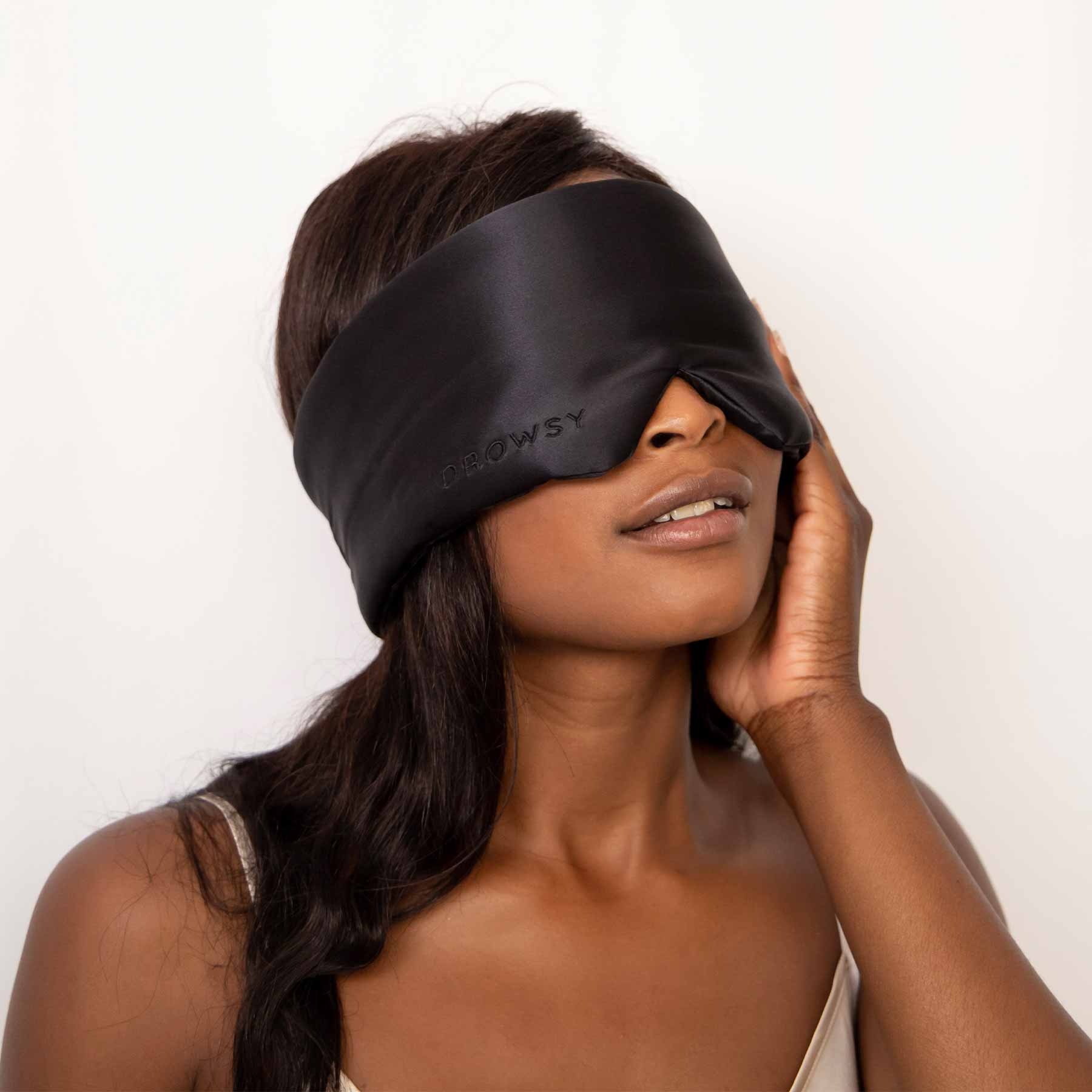 A woman with long dark hair, in a light top, gently touches her face while wearing the drowsysleepco 8 Hrs Deep Sleep Sets black silk mask. She is posed against a plain white background.