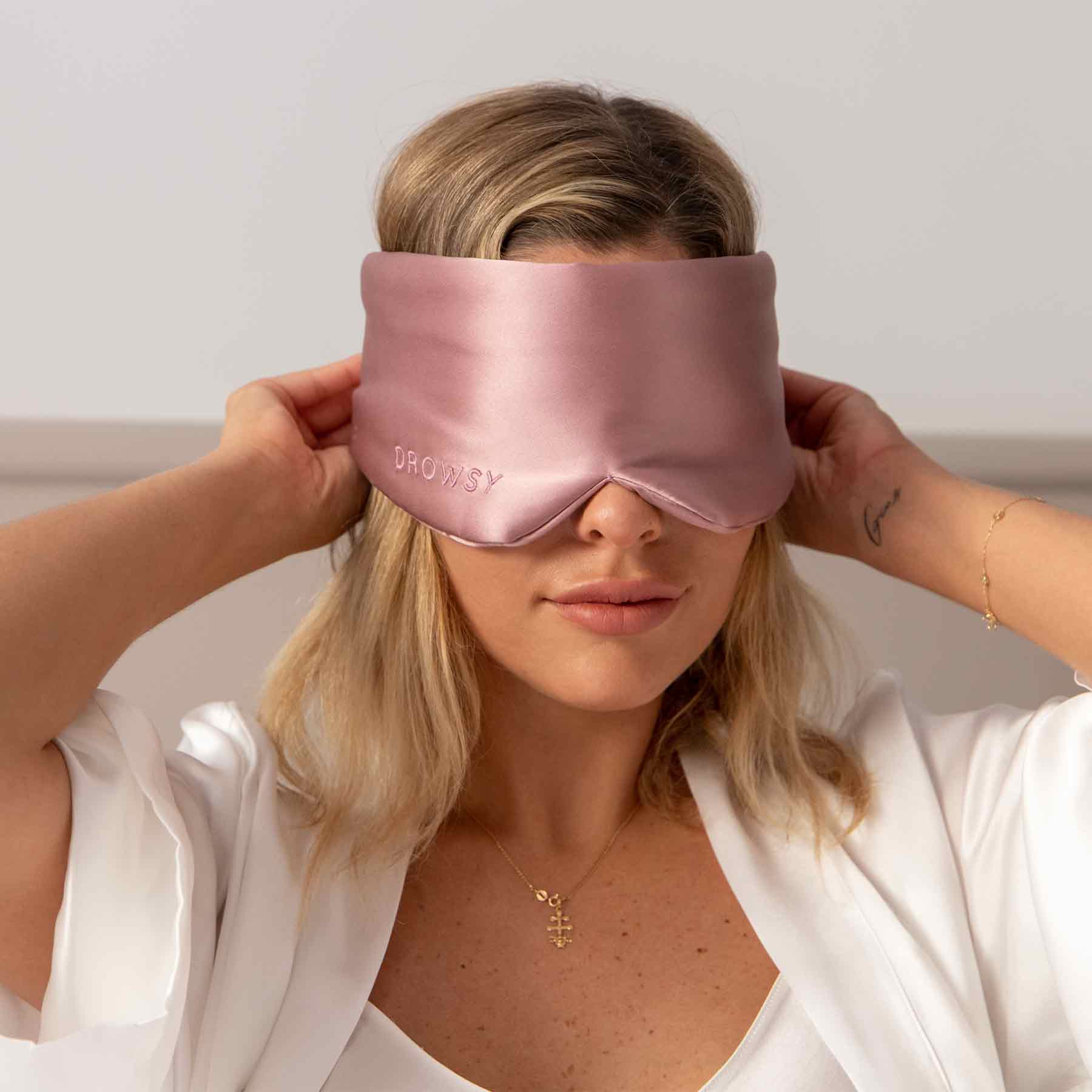 A blonde woman in a white robe wears a silky Damask Rose sleep mask by drowsysleepco, touching its sides. The mulberry silk eye mask features the word DROWSY embroidered on it.