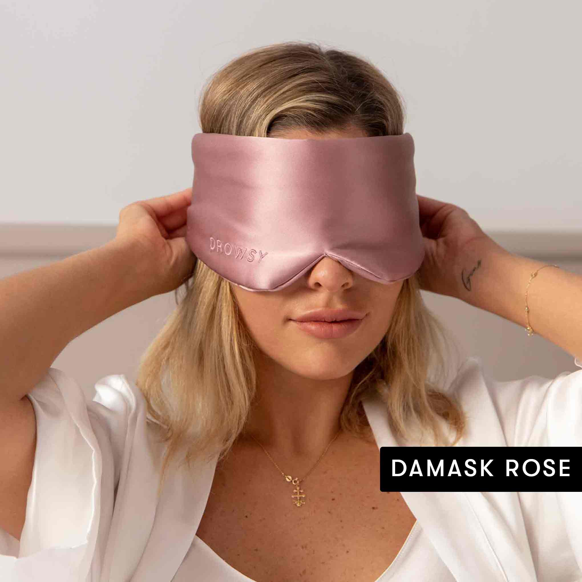 A woman in a white robe and gold cross necklace adjusts a light pink drowsysleepco Lovers Pack silk sleep mask over her eyes. DAMASK ROSE appears in the corner.