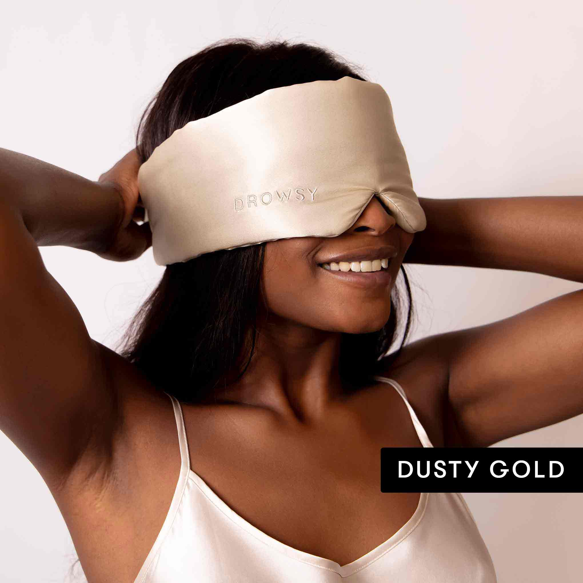 A smiling woman adjusts a drowsysleepco Lovers Pack luxury mulberry silk blackout eye mask over her eyes. She wears a matching satin top, and her long dark hair cascades down. The words DUSTY GOLD appear in a black box.