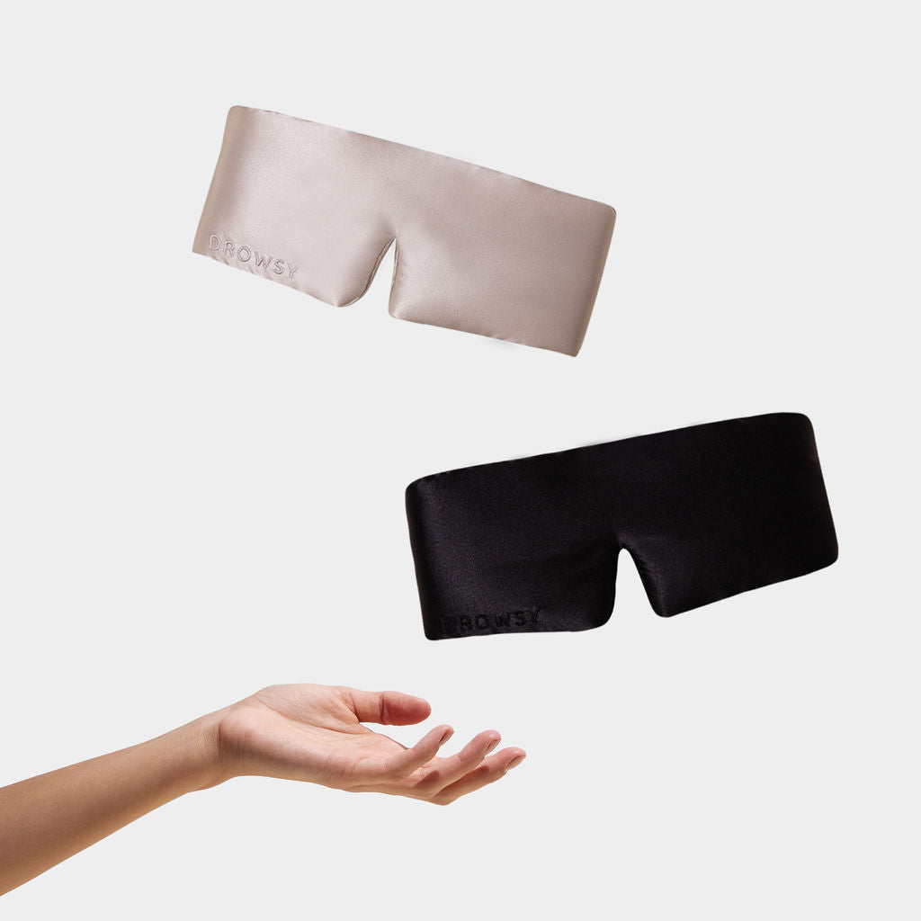 A hand tosses two drowsysleepco Lovers Pack blackout eye masks, one beige and one black, made from luxury mulberry silk, both featuring “BROWSY,” against a plain white background.