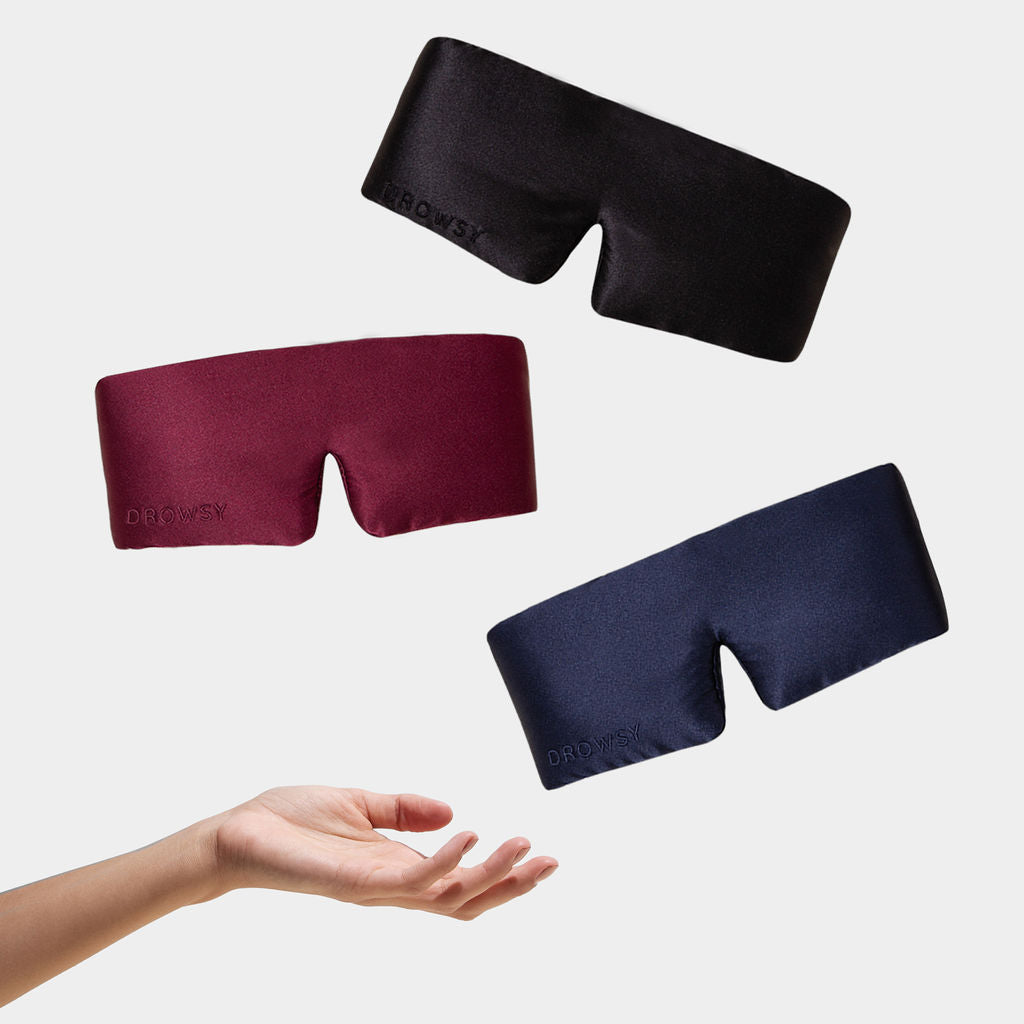 The drowsysleepco Besties Pack, featuring three luxury silk sleep masks in black, burgundy, and navy blue, hovers above an outstretched hand on a simple white background.