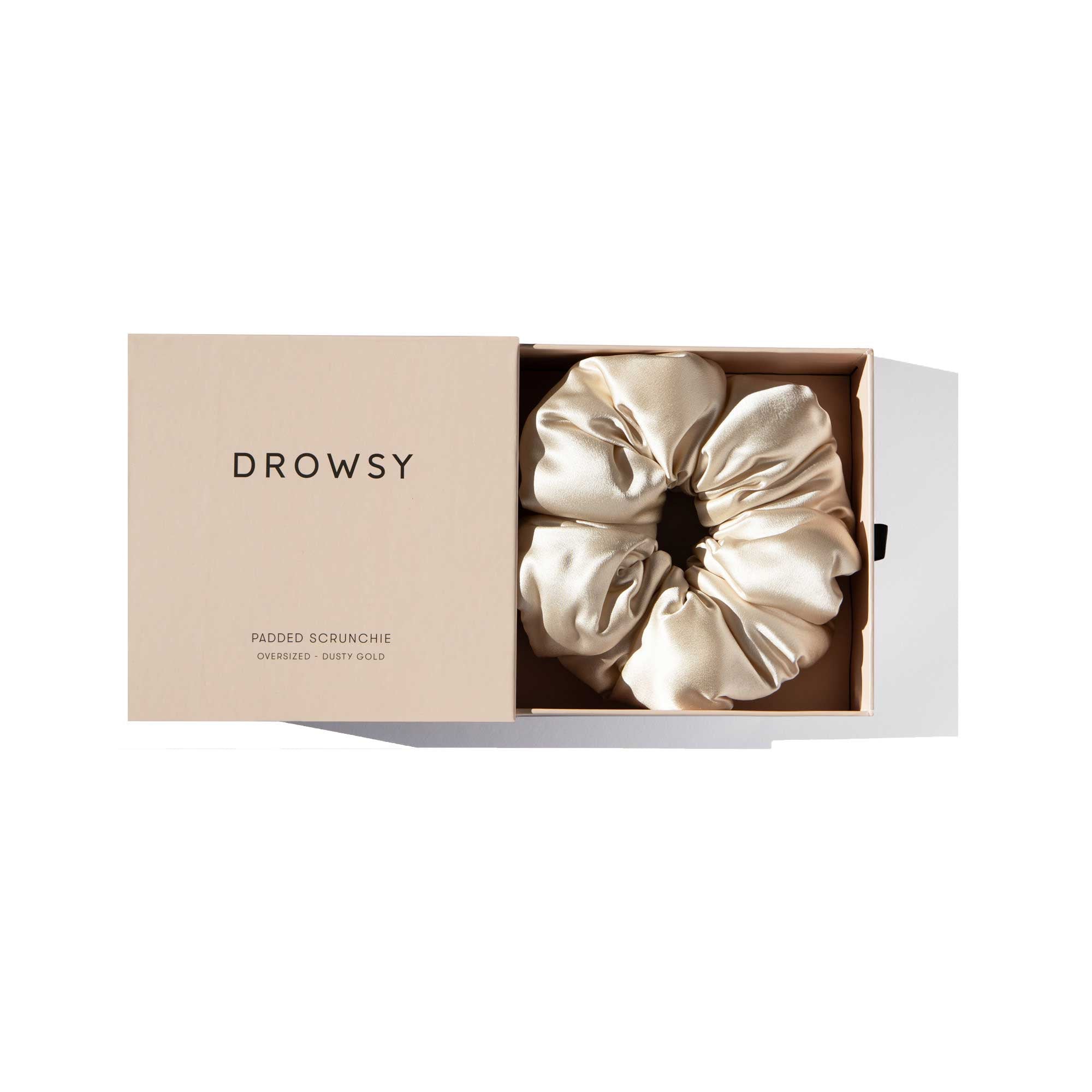 A beige box labeled "drowsysleepco" with the lid partially open reveals a large, shiny, light gold Rajah Padded Scrunchie neatly placed inside.