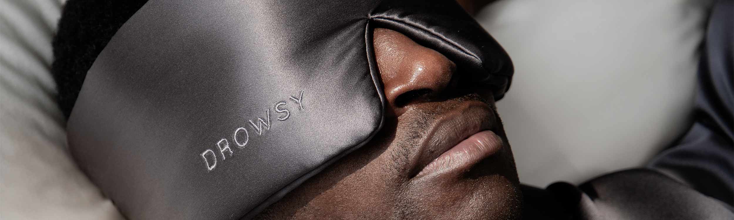 A person wearing a large, silky black sleep mask labeled DROWSY rests their head on a pillow, suggesting relaxation and sleep.