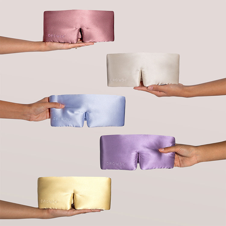 Five hands each hold a silk sleep mask in different colors—pink, cream, blue, lavender, and yellow—against a plain light background. Each mask has a small nose cutout and embroidered logo.