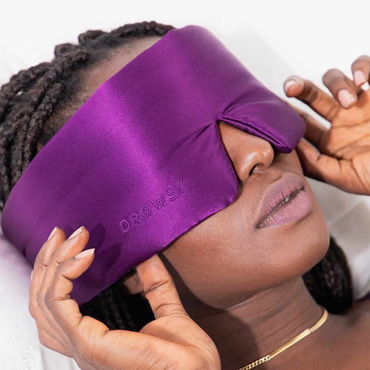 A person with braided hair lies down, relaxing as they adjust a purple silk Sleep Mask by drowsysleepco, embroidered with “DROWSY.”.