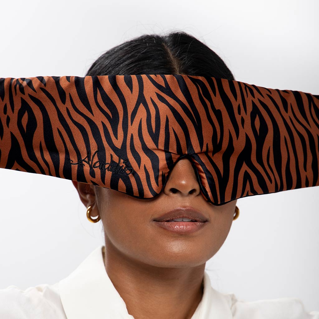 A woman with gold hoop earrings and a white collared shirt wears the drowsysleepco Rajah Sleep Mask, featuring a wide brown and black tiger-striped mulberry silk design, covering her eyes and most of her forehead.