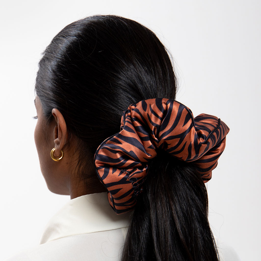 A woman with long, dark hair pulled back in a low ponytail wears the drowsysleepco Rajah Padded Scrunchie, featuring a bold black and orange tiger stripe pattern, styled with a white top and gold hoops for chic hair protection.