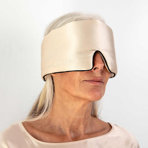 An older woman with long gray hair wears the Dusty Gold - Pipe Dreams Edition sleep mask by drowsysleepco over her eyes. She looks calm, dressed in a matching satin top, against a plain white background.
