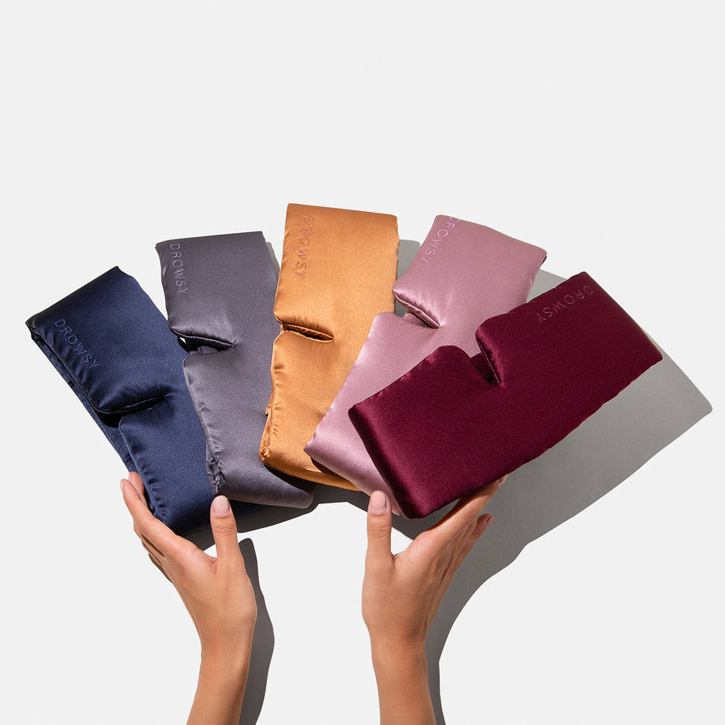 Two hands hold the drowsysleepco Family Pack: five luxury silk masks in navy blue, gray, gold, pink, and burgundy. Each folded mask features a subtle embroidered logo—ideal for beauty sleep—displayed against a plain white background.