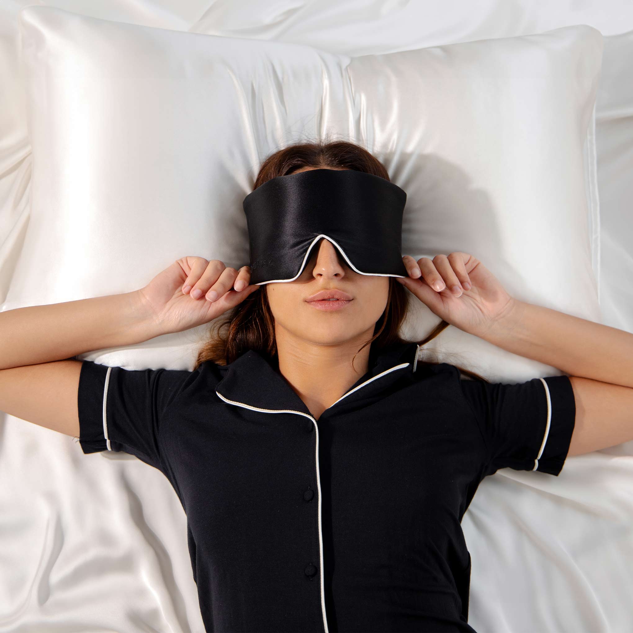 A woman lies on a white silk bed, adjusting a black sleep mask. Seen from above, she's surrounded by smooth bedding featuring the Jasmine Cushion Cover from drowsysleepco.
