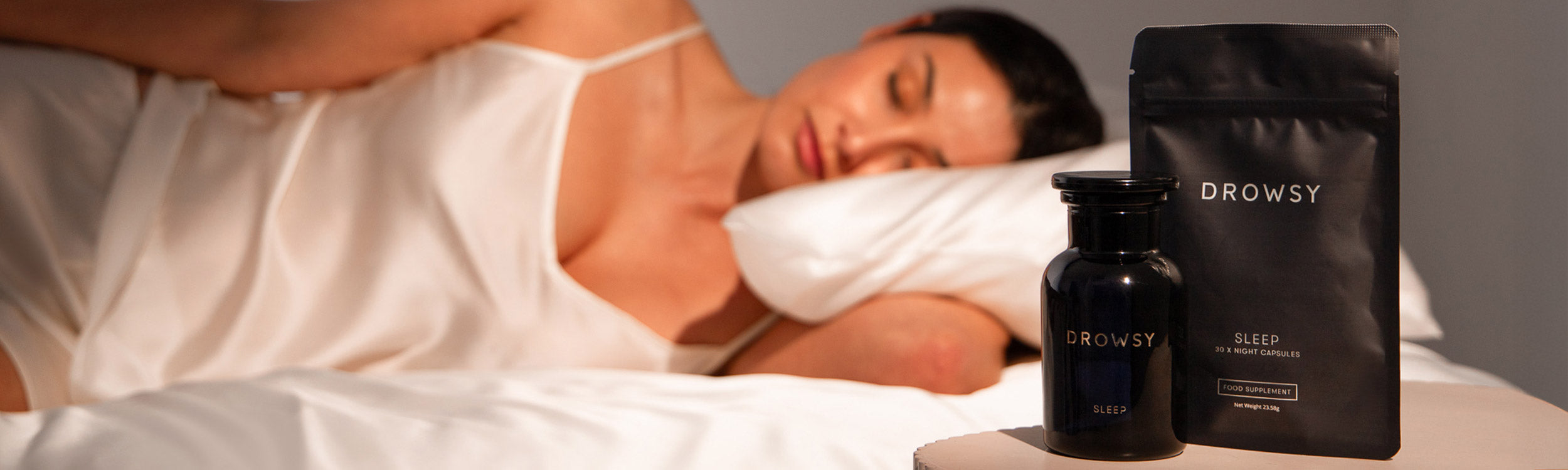 A woman in a white satin nightgown sleeps on a bed. In the foreground, there are two Drowsy-branded sleep products: a bottle and a packet labeled “Sleep,” placed on a bedside table.