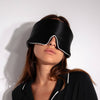 A woman with long brown hair wears the Black Jade - Pipe Dreams Edition sleep mask by drowsysleepco, covering her eyes. She pairs it with a matching black top featuring white piping, standing before a plain white backdrop.