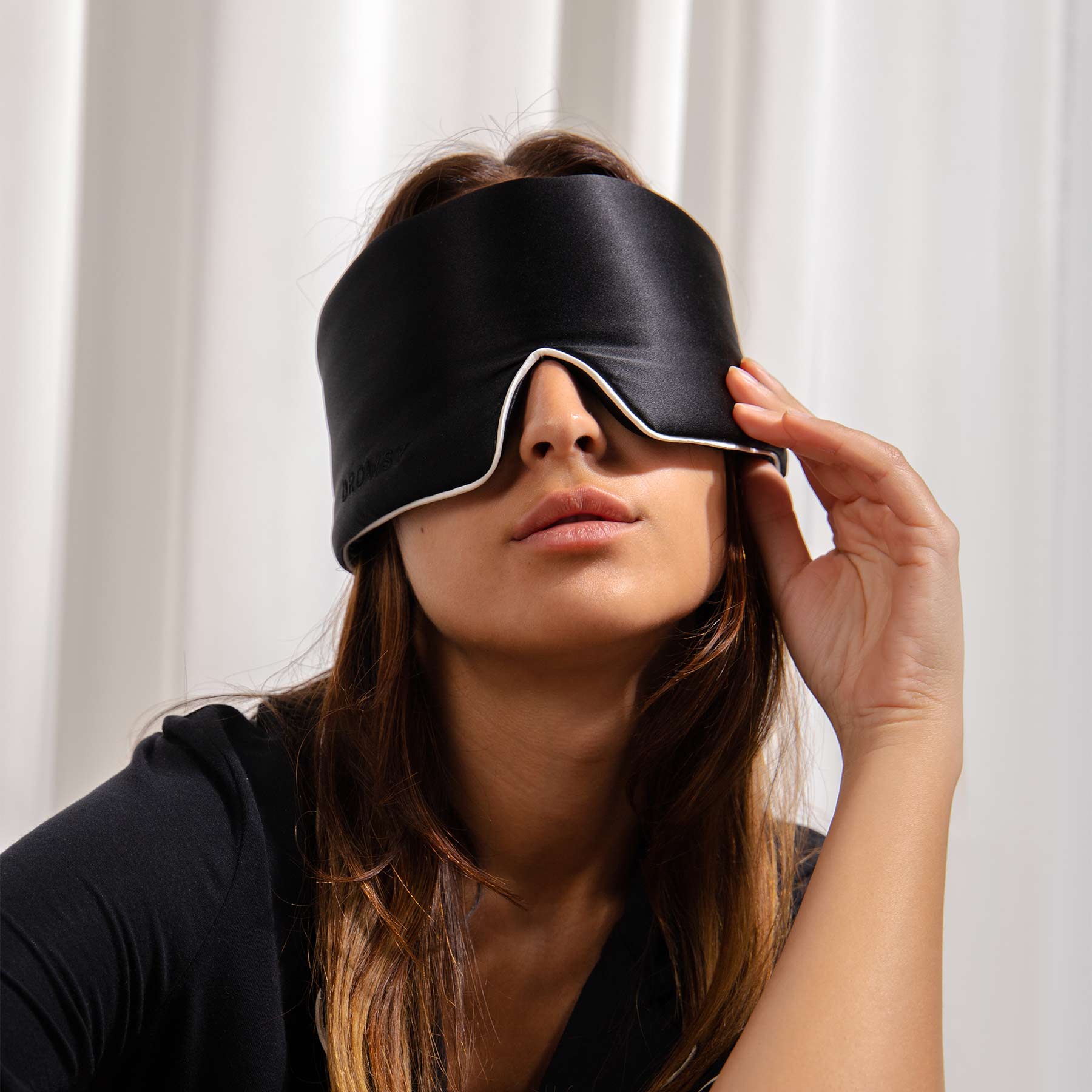 A woman with long brown hair, wearing the drowsysleepco Black Jade - Pipe Dreams Edition sleep mask and a black shirt, holds the edge of the mask as she sits in front of white curtains.