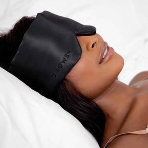 A woman relaxes on a white bed, wearing the drowsysleepco Perfect Escape Bundle - Black Jade silk sleep mask with "Drowsy" embroidery, enjoying restful beauty sleep as her head rests on a pillow.