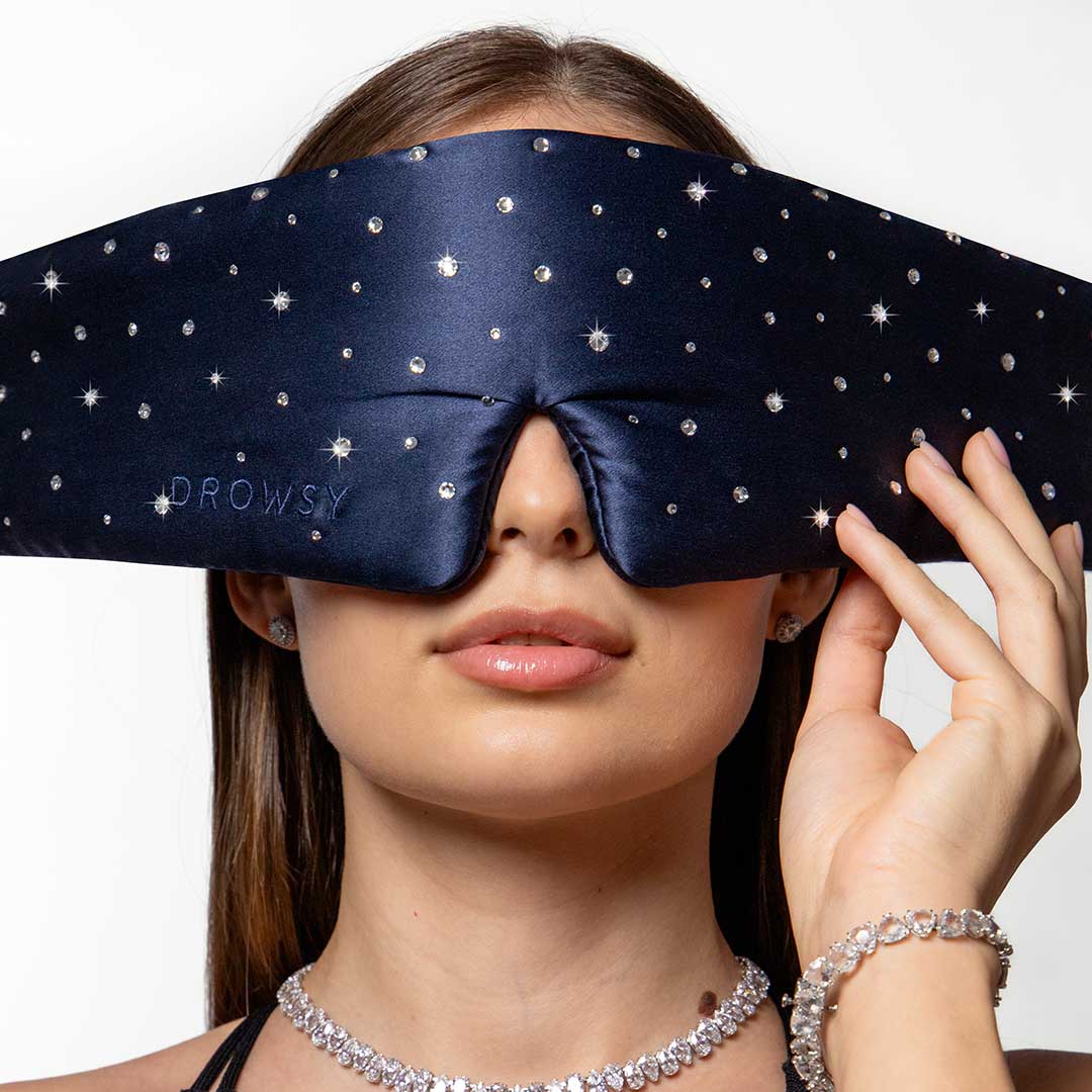 A woman wearing a dark blue, star-patterned sleep mask that covers her eyes and eyebrows, touching the mask with one hand. She is accessorized with a sparkling necklace, bracelet, and earrings.