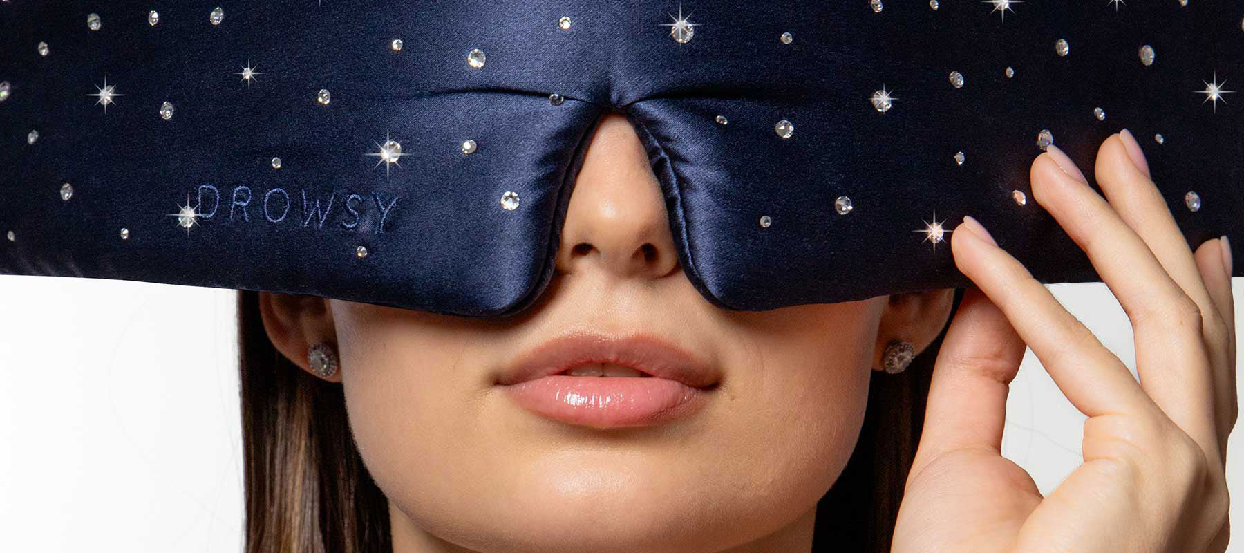 A woman wears a navy blue, rhinestone-studded sleep mask that covers her eyes and eyebrows. The mask is embroidered with the word DROWSY. She gently holds the edge of the mask with one hand.