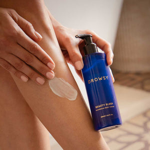 As part of their night ritual, a person applies a white cream from a blue bottle labeled drowsysleepco Beauty Sleep Body Cream to their leg.