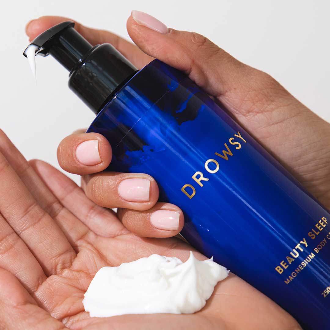 A hand with light pink manicured nails dispenses white Beauty Sleep Body Cream by drowsysleepco from a blue bottle onto the palm—perfect for your nighttime routine.