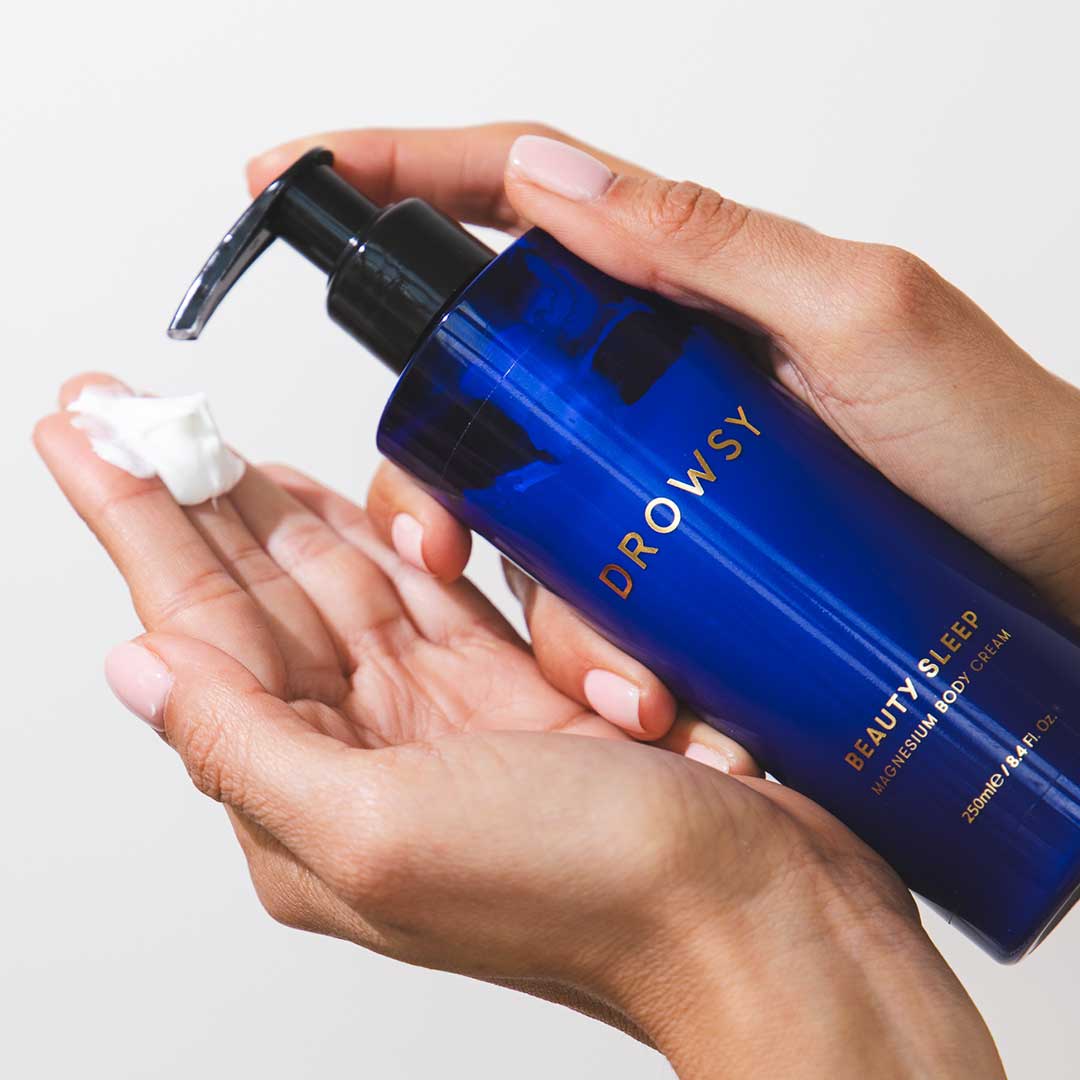 A person holds a blue bottle labeled Beauty Sleep Ritual by drowsysleepco, dispensing white cream onto their palm as part of their magnesium body care routine.