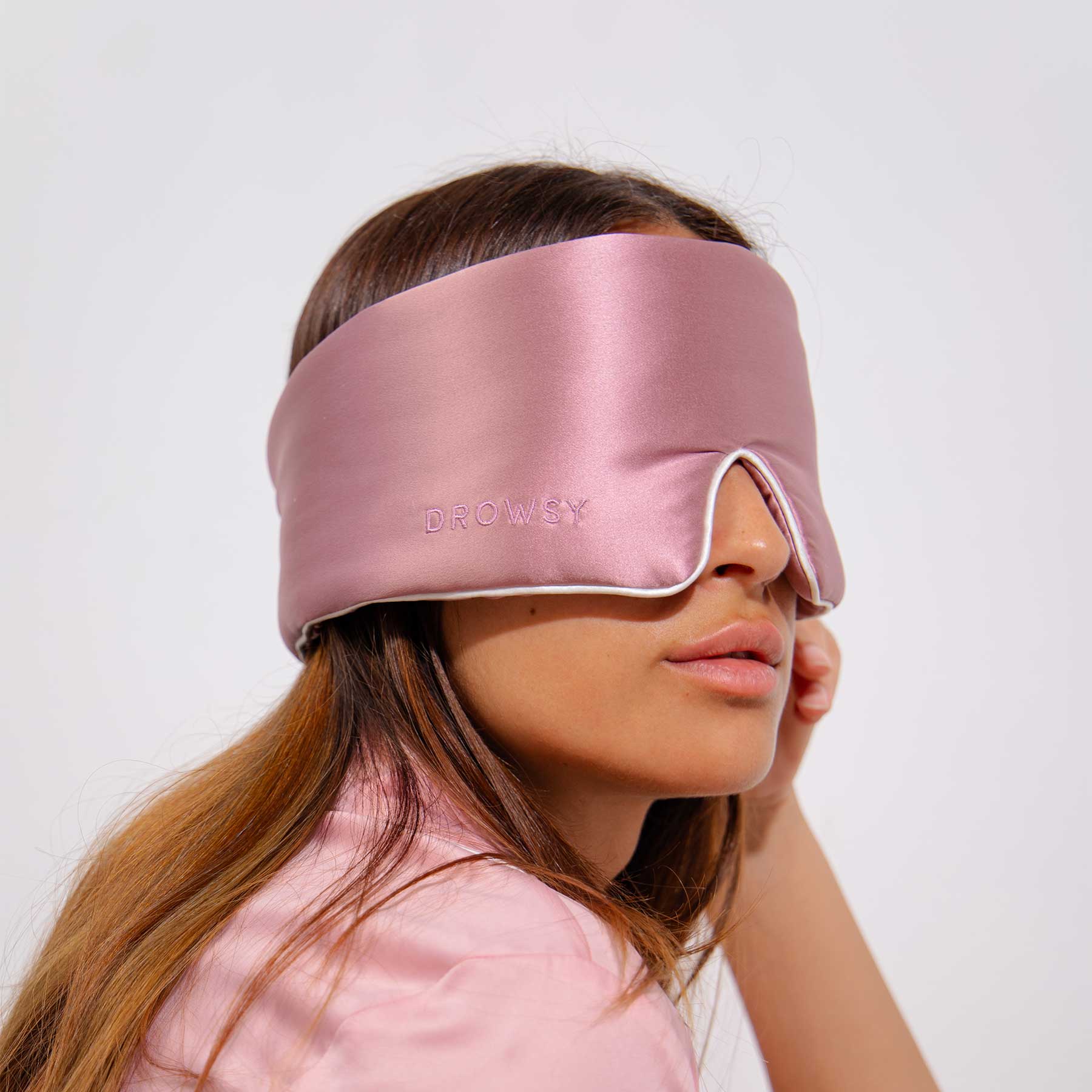 A woman with long brown hair wears the Damask Rose - Pipe Dreams Edition sleep mask by drowsysleepco over her eyes. The shiny pink adjustable mask matches her pink top as she poses against a plain white background.