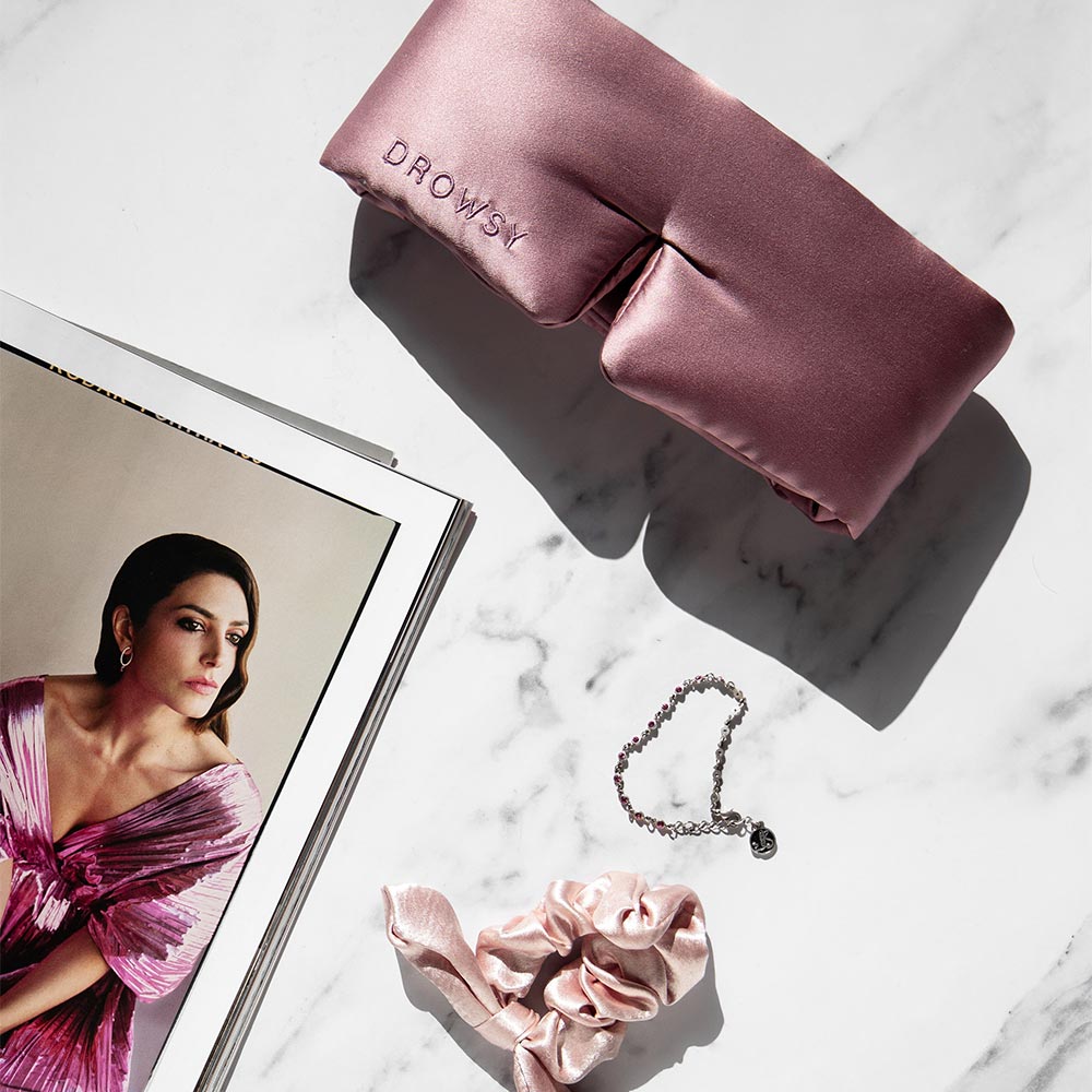 The Damask Rose sleep mask and matching scrunchie by drowsysleepco are displayed with a silver bracelet and an open magazine showing a woman in pink, all arranged on a white marble surface.