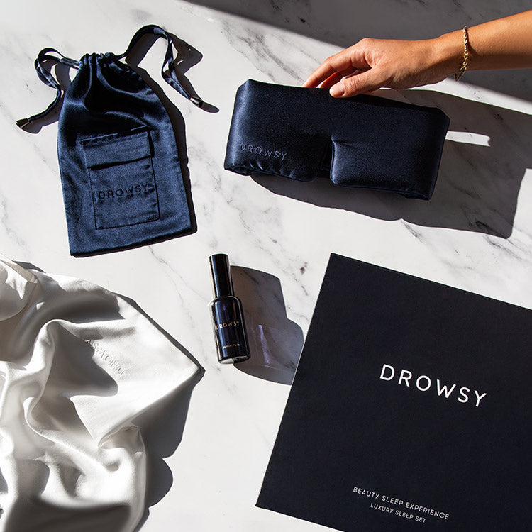 A person’s hand holds a black satin sleep mask labeled “Drowsy” on a marble surface, alongside a matching drawstring pouch, a spray bottle, a white satin cloth, and a black box with “Drowsy” branding.