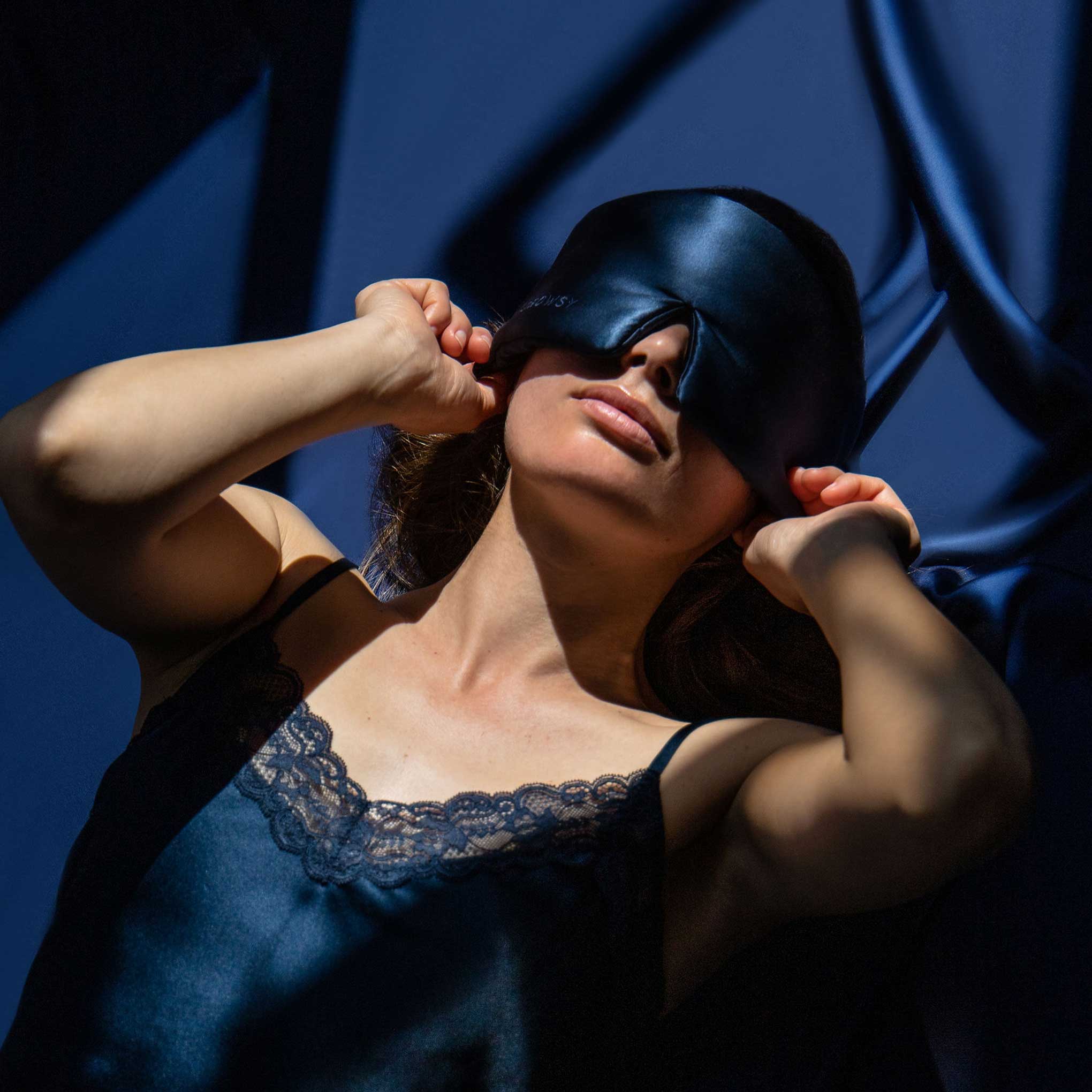 A woman wearing a navy satin slip dress and a matching satin sleep mask adjusts the mask over her eyes. She is lit by dramatic sunlight against a dark blue satin background.