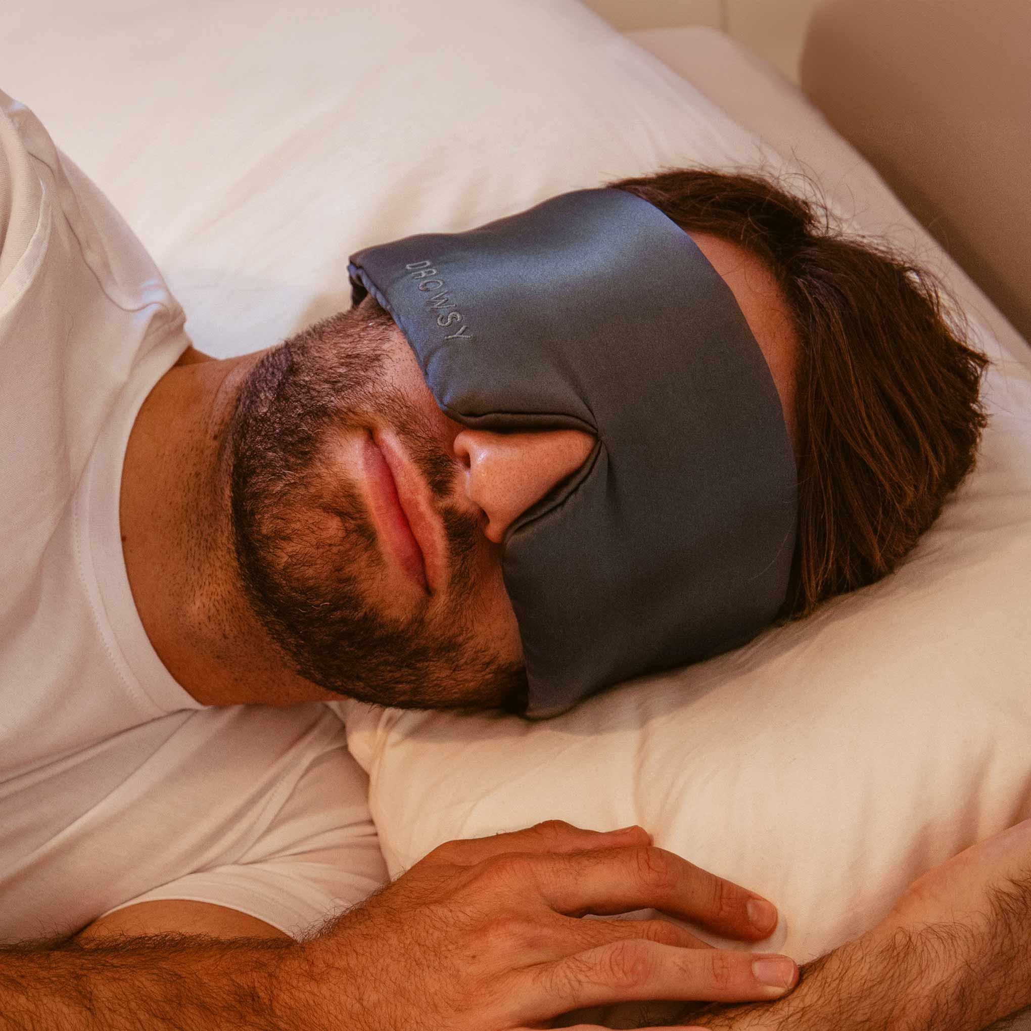 A man with a beard rests on a pillow, wearing a drowsysleepco 2-Pack Sleep Mask in Midnight Blue. He is dressed in white and has one hand on the bed.